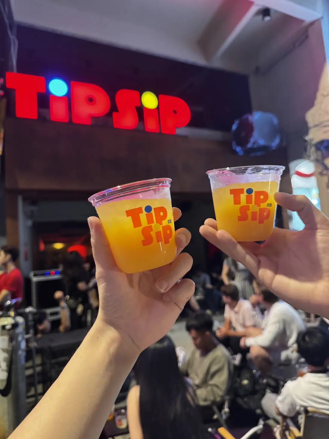 Two TipSip cups raised in a toast under the TIPSIP neon sign
