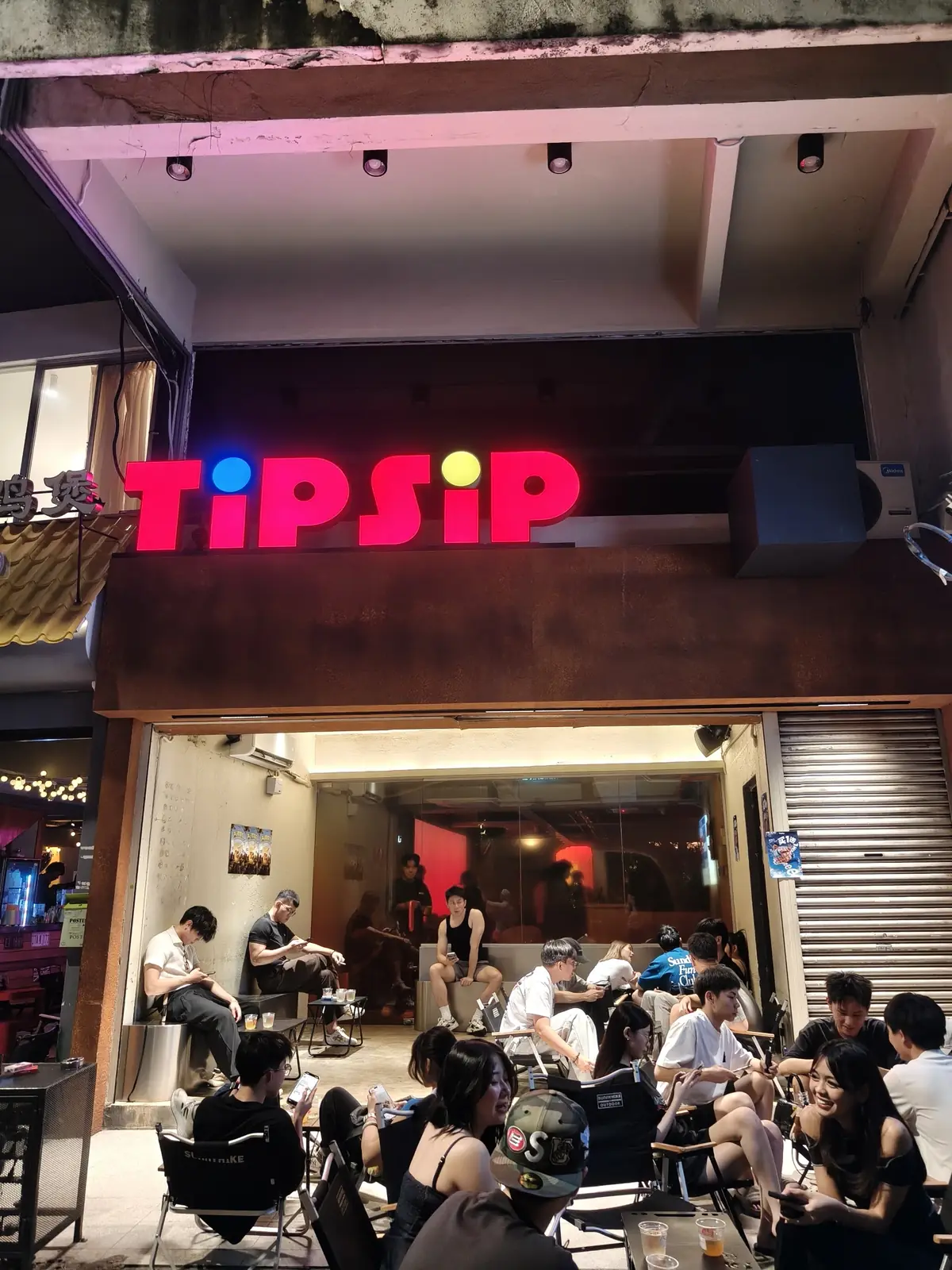 TipSip storefront at night with the red TIPSIP sign and a crowd on camping chairs